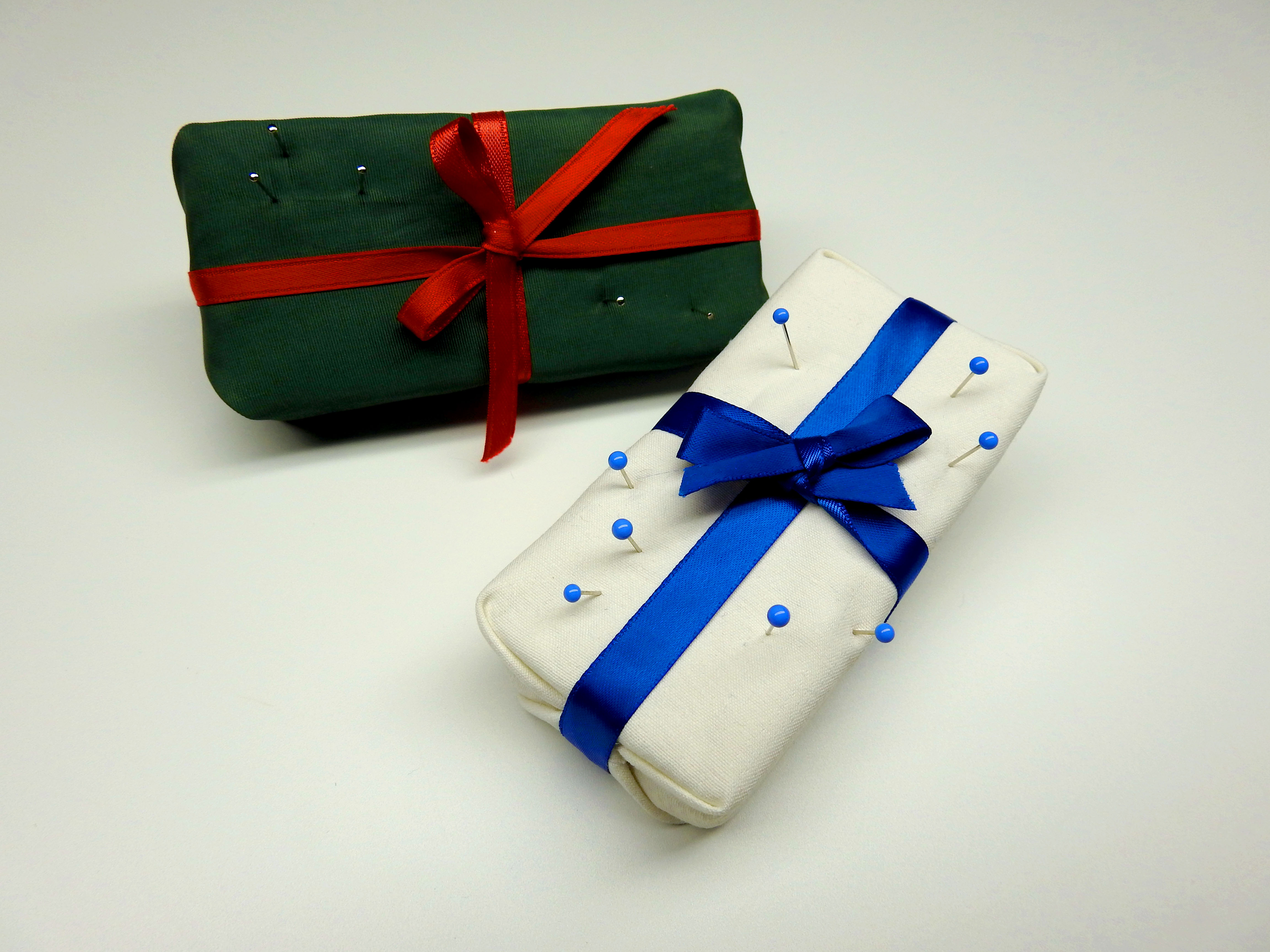 Two finished gift box pin cushions with pins stuck in them. One is white with a deep blue ribbon (winter,) the other is green with a red ribbon (joyful.)